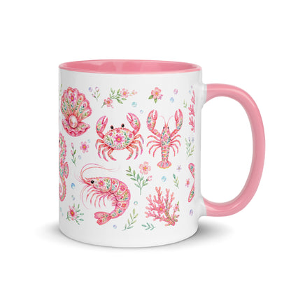 Weiße Keramiktasse mit rosa Henkel und Innenrand sowie verspieltem Muster aus Krabben, Hummern, Muscheln und Blumen.